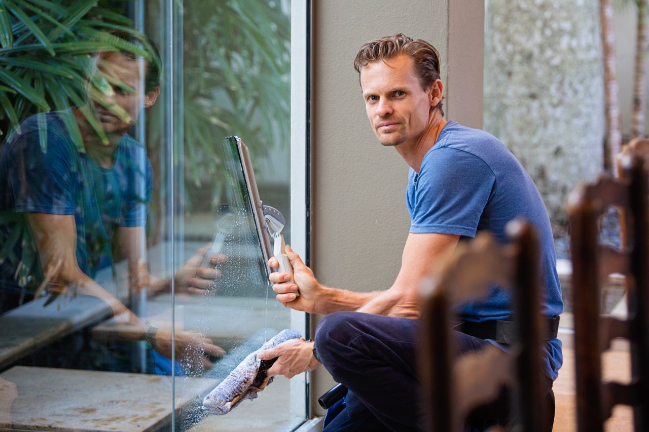 services-01 A man in a blue t-shirt cleaning large glass pane with a squeegee indoors.
