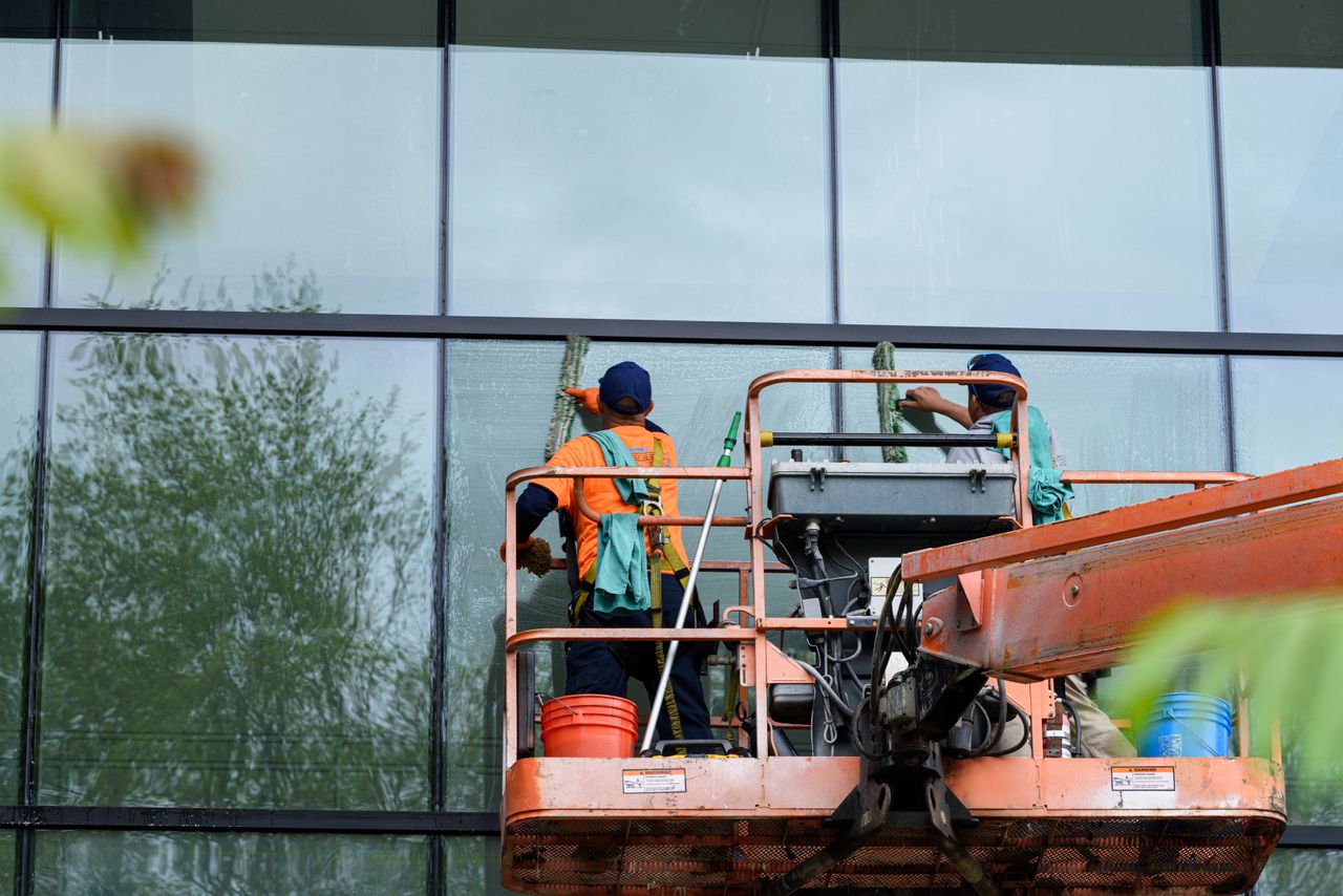 services-04 Two workers cleaning windows on a glass building facade using a lift platform, enhancing building aesthetics.